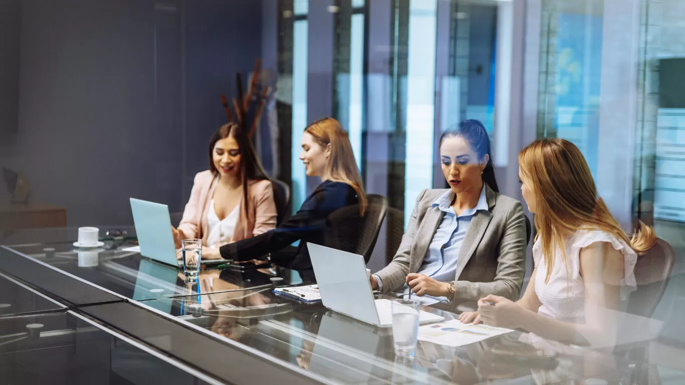 Four professional women collaborating in a modern conference room, using laptops and discussing documents, emphasizing teamwork and technology solutions for CPA firms.