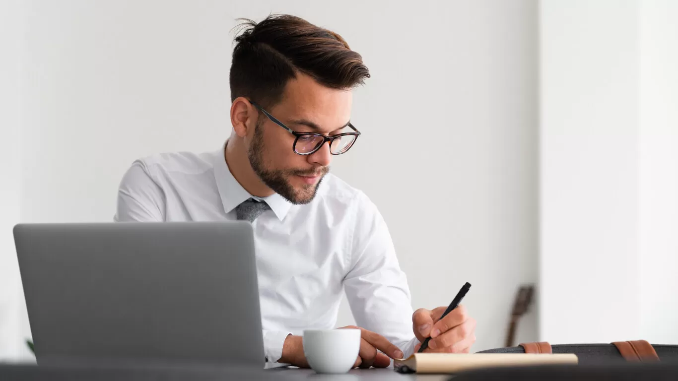 Man in a white shirt and glasses writing notes while working on a laptop, illustrating productivity and efficiency for CPA firms using PracticeERP technology solutions.