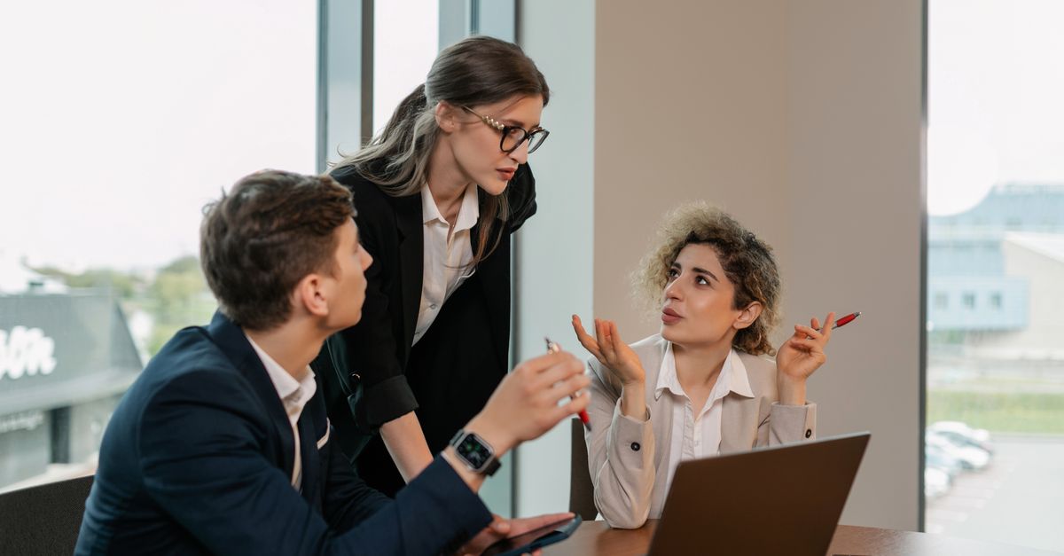 Three professionals in a business meeting, discussing work around a laptop in a modern office.