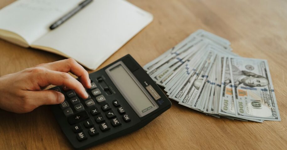Close-up of a hand using a calculator with cash and a notebook on a wooden table.