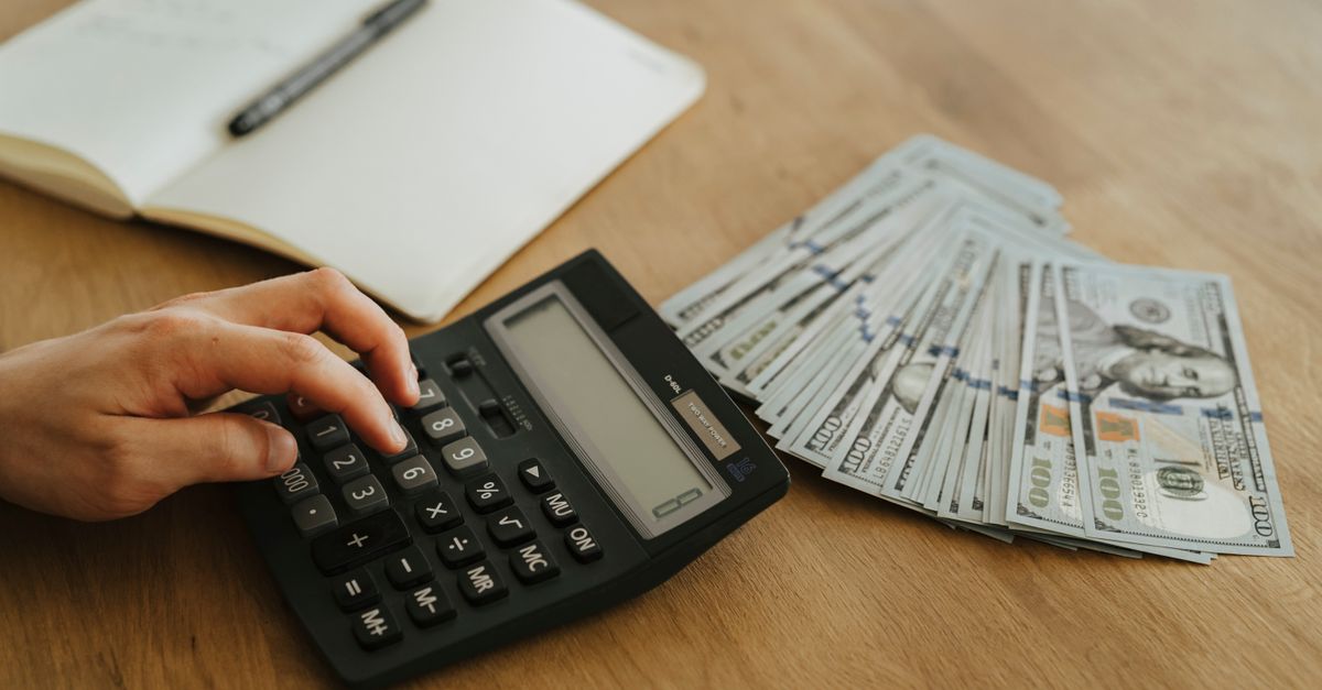 Close-up of a hand using a calculator with cash and a notebook on a wooden table.