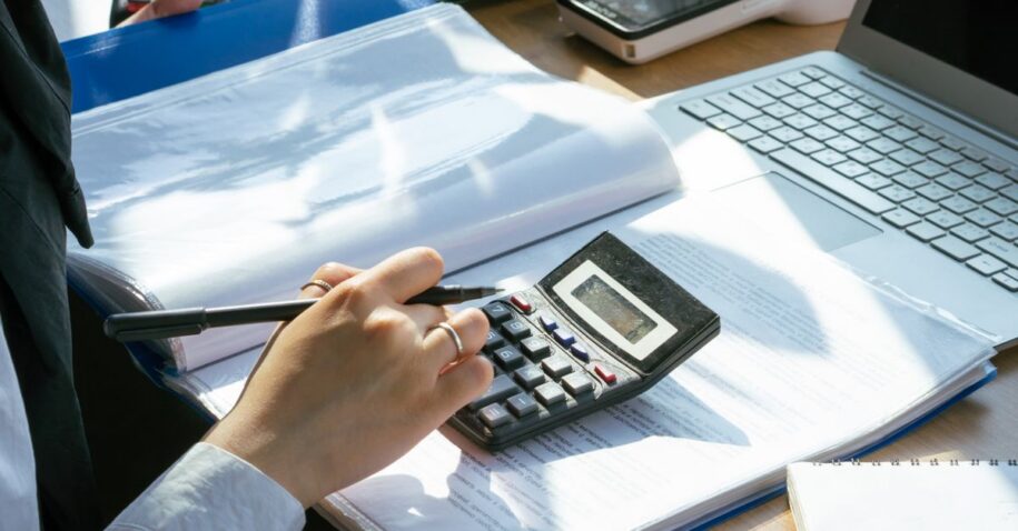 A person working on financial calculations using a calculator and laptop at an office desk.