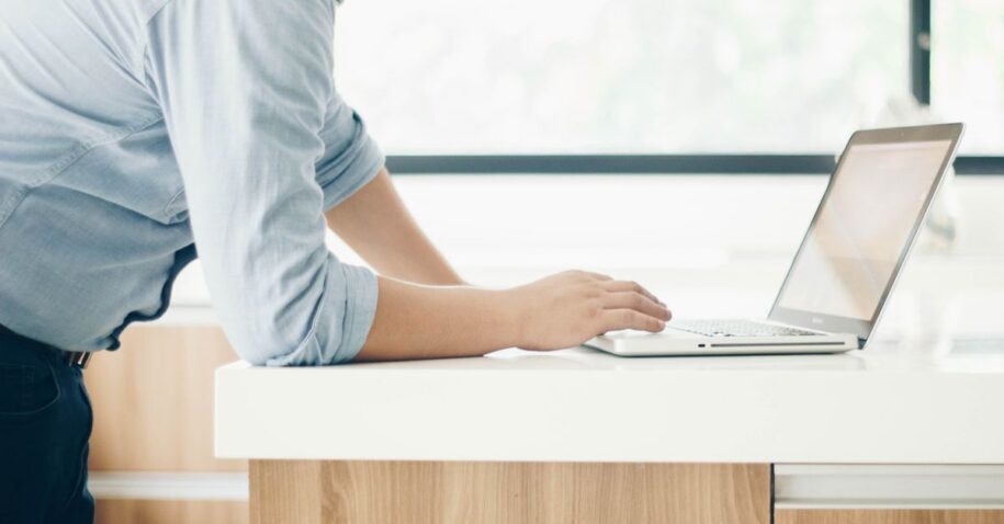 A man leaning on a desk working on a laptop in a bright indoor office setting.