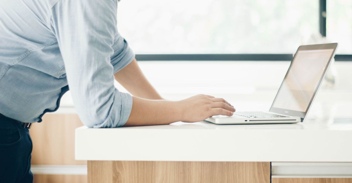 A man leaning on a desk working on a laptop in a bright indoor office setting.
