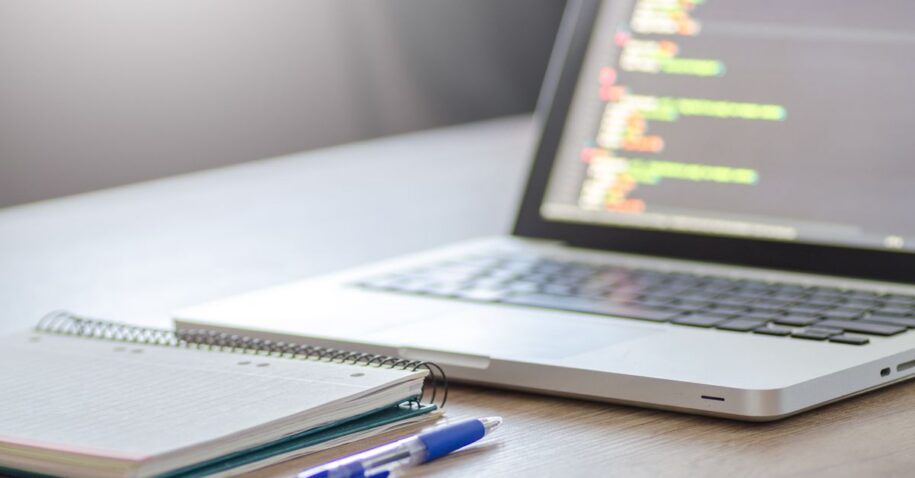 Open laptop with programming code on screen next to a notebook and pen on a desk.