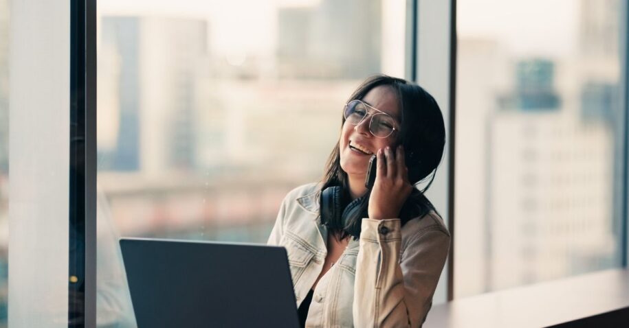 A joyful woman with glasses and headphones chats on phone by laptop in a modern office.