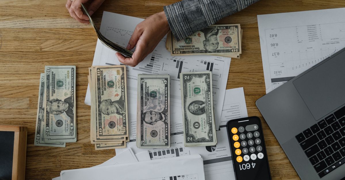 Person counting cash with a calculator and documents on a wooden desk, representing financial planning.