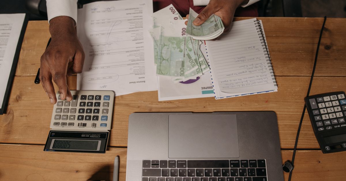 Close-up of a person counting cash with documents and a laptop in an office setting.