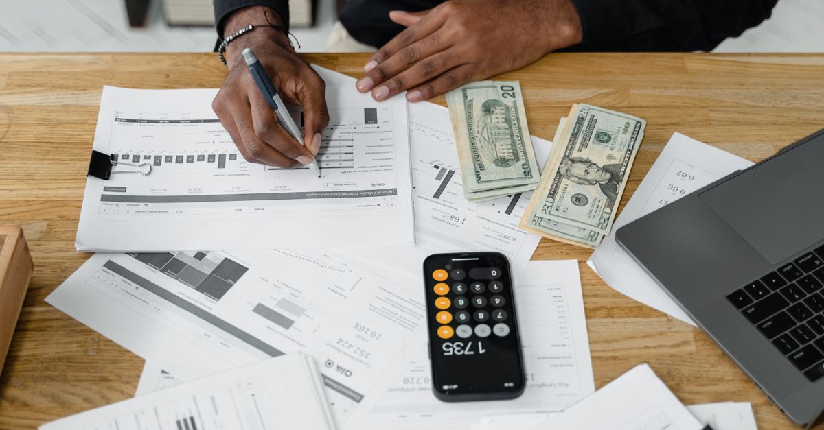 Man working on financial reports with calculator, money, and laptop on a desk.