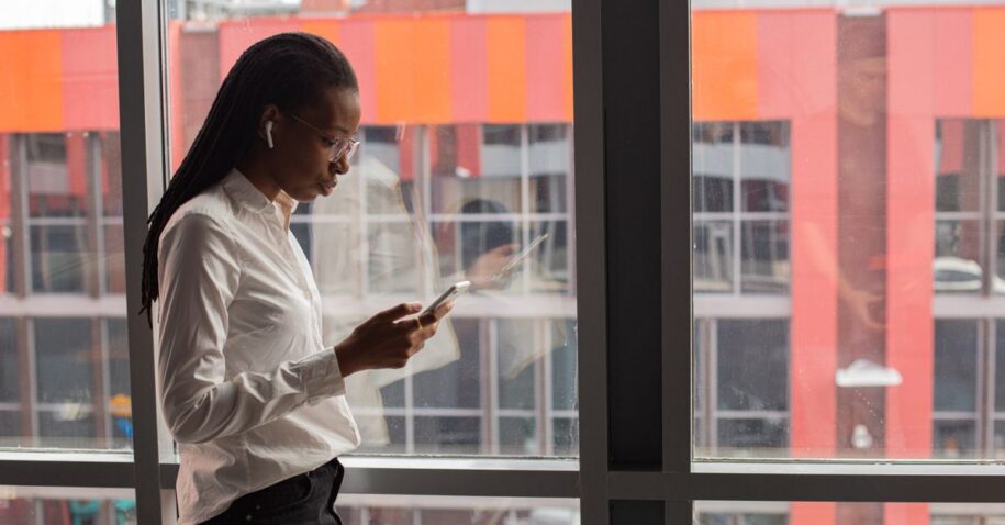 African American woman focused on her smartphone by a large office window, urban background.