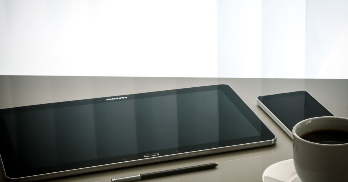 Minimalist office desk setup featuring a tablet, smartphone, pen, and coffee cup on a reflective surface.