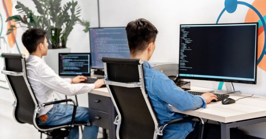 Two male developers at desks programming in a modern office workspace with large monitors.