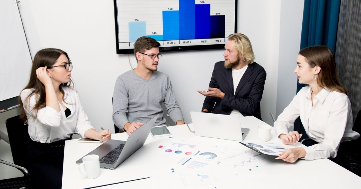 A group of business professionals discussing data at a meeting table with laptops and charts.