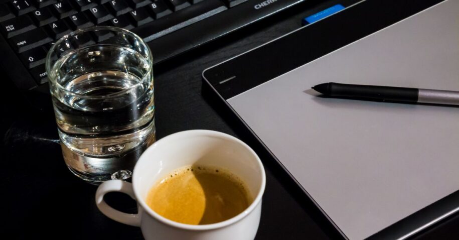Office desk setup with a coffee cup, glass of water, tablet, pen, and keyboard.
