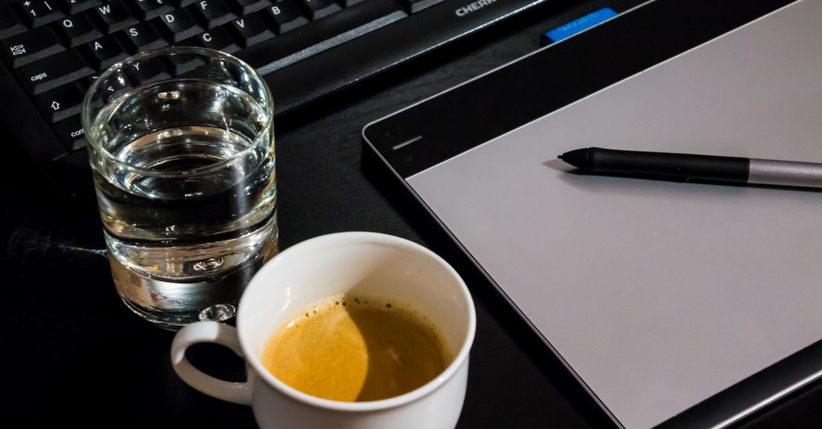Office desk setup with a coffee cup, glass of water, tablet, pen, and keyboard.
