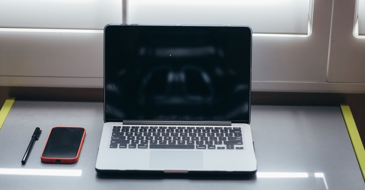 A minimalist desk setup featuring a laptop, smartphone, and pen under soft window light.