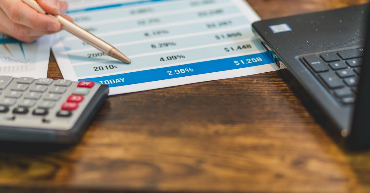 Close-up of a hand with pen analyzing financial rates on paper with a calculator and laptop nearby.